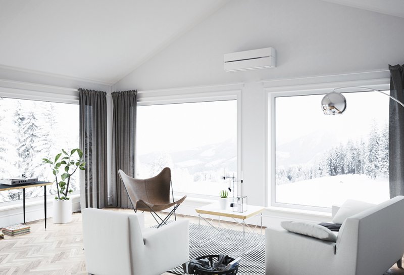 Hvit varmepumpe montert under himling i stor, lys stue med vinterlandskap utenfor.