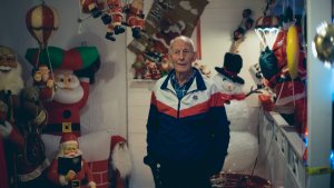 Tore Karlsen står ved inngangspartier til huset med en tynn sportsjakke på seg. I bakgrunnen ser man nisser, snømenn og juletrær i figurer som er hengt opp på veggen og lyser.