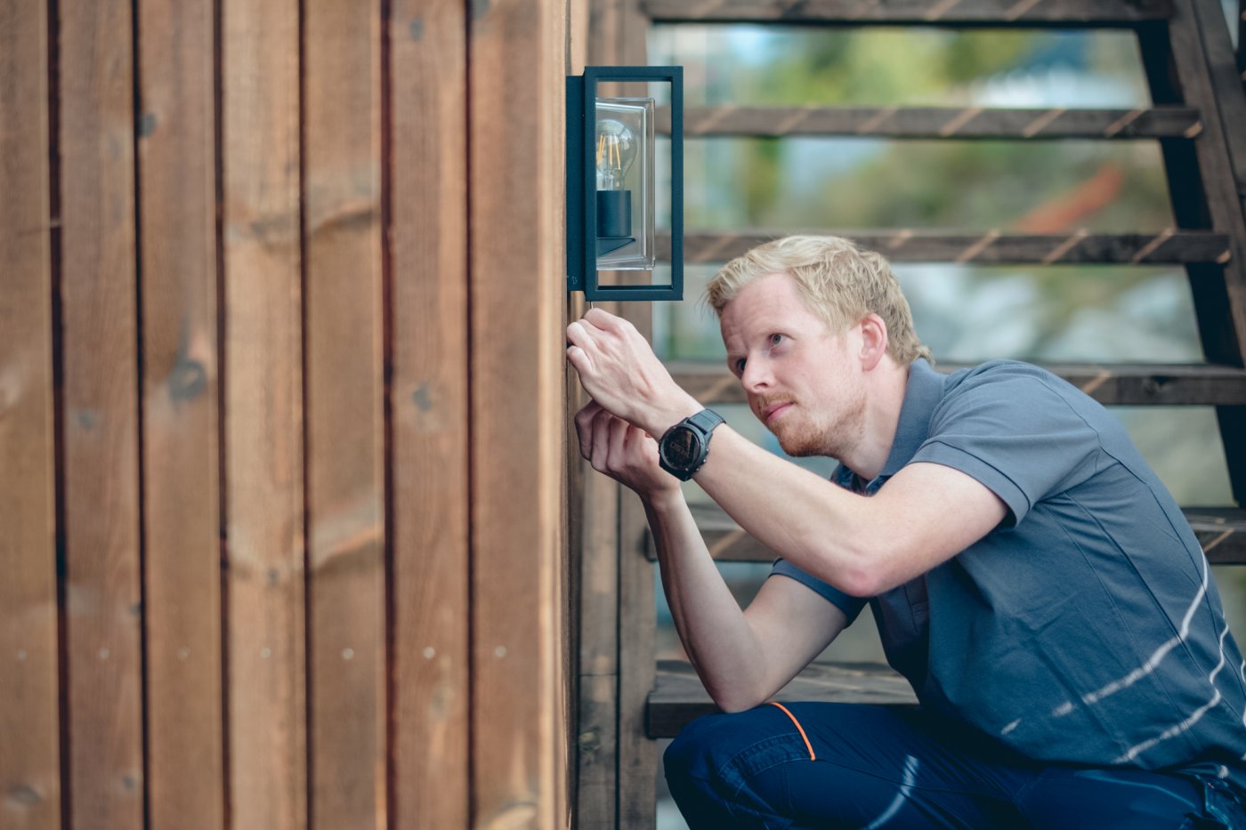 Ole-Magnus sitter på huk og lener seg inn mot ytterveggen av huset mens han skrur opp en utelampe på veggen.