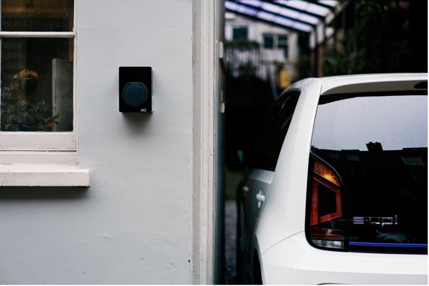 Verdens minste elbillader EO Mini fra Micro Matic på grå husvegg i mur med hvit elbil parkert ved siden av.