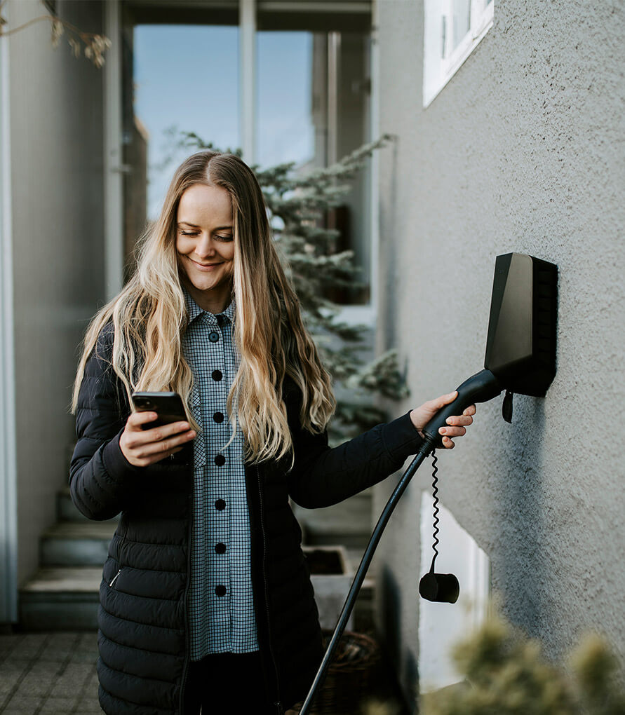 Dame med langt lyst hår. Hun ser på mobilen samtidig som hun holder den andre hånden på ladekabel til bil.