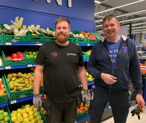 To menn som står smilende foran fruktavdelingen på Rema1000.